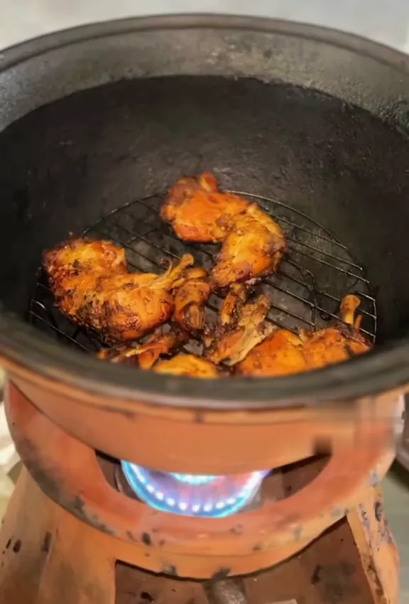 Ayam di atas jaring dalam tungku gerabah, api kompor biru menyala di bawahnya